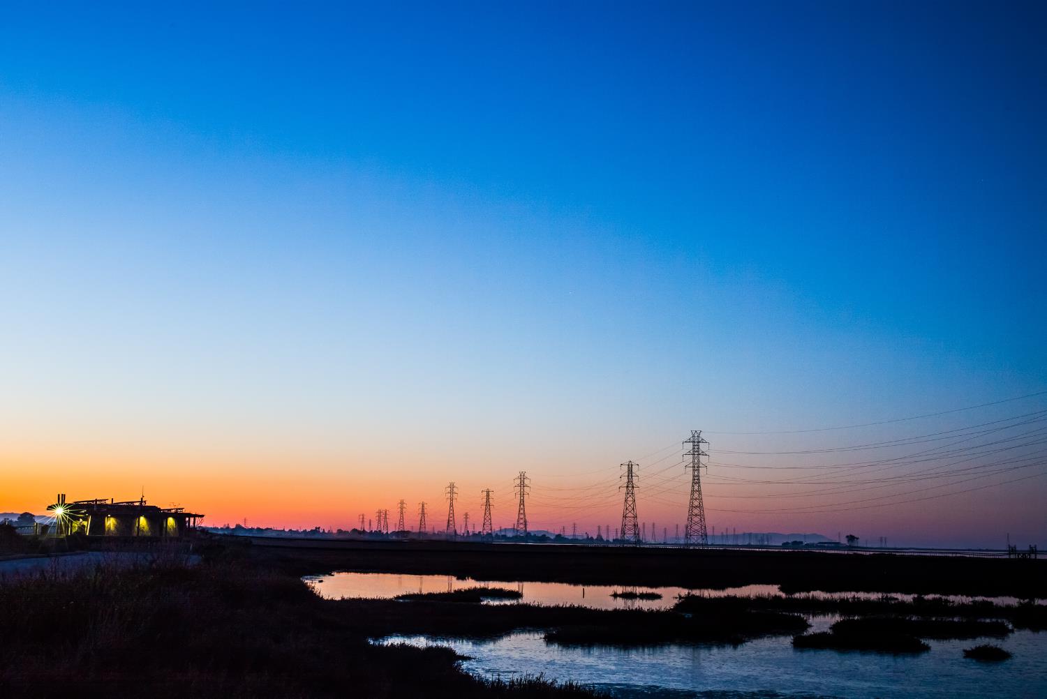 A colorful orange-yellow sunset view of the Baylands with the lighted interpretive center and distant electrical towers silhouetted against the darkening sky