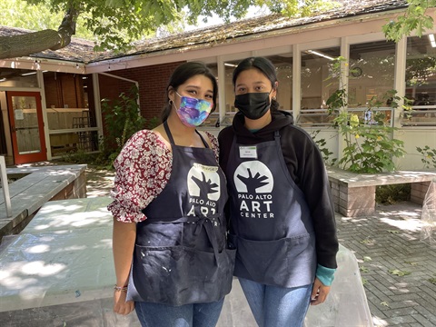 Two people stand at the Palo Alto Art Center wearing aprons and masks