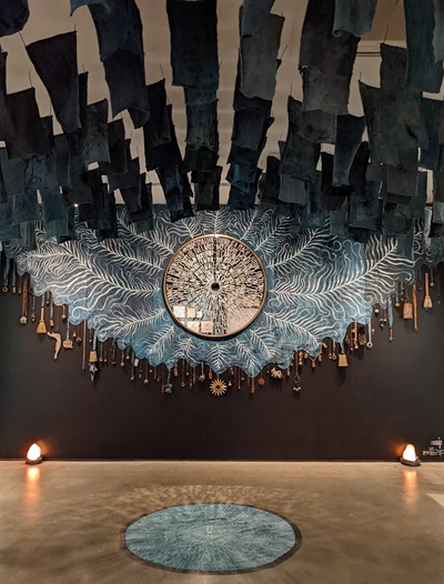 Installation image of Drift, with circular mirror on wall,circular pattern on floor, blue fabric strips hanging from the ceiling and found objects hanging from blue fabric on wall