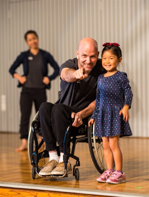 Performance by Axis Dance at the Art Center, a little girl has been asked to go up to the stage, she stands with a wheelchair using dancer who points at the camera